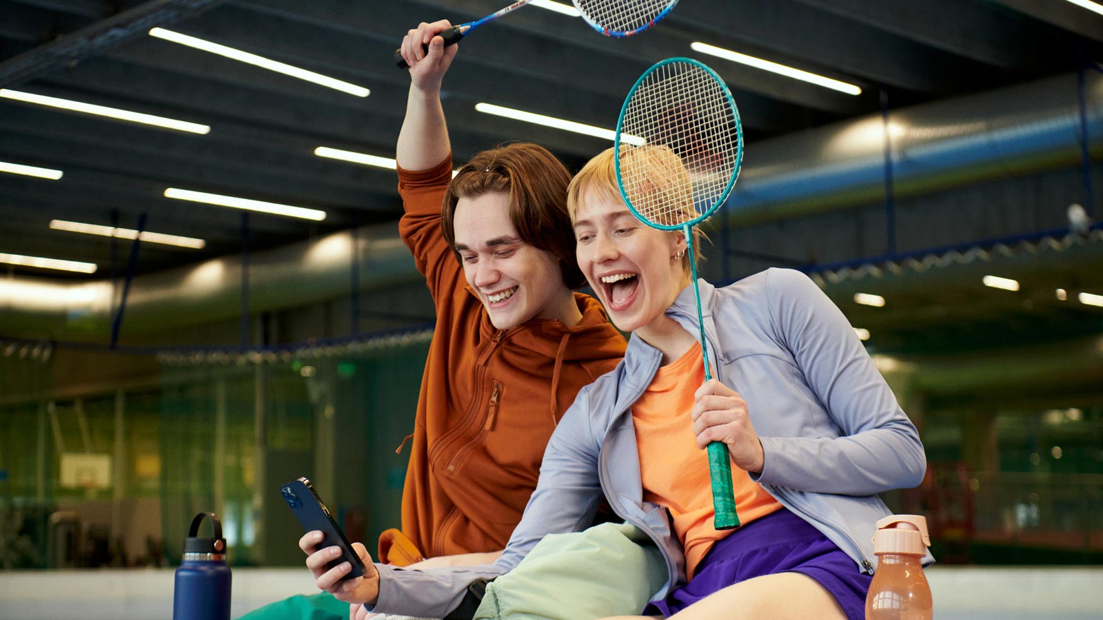 Kaksi henkilöä urheiluhallissa, jotka istuvat tauolla sulkapallokentän laidalla. Heillä on käsissään sulkapallomailat ja he katsovat iloisina kännykkää.