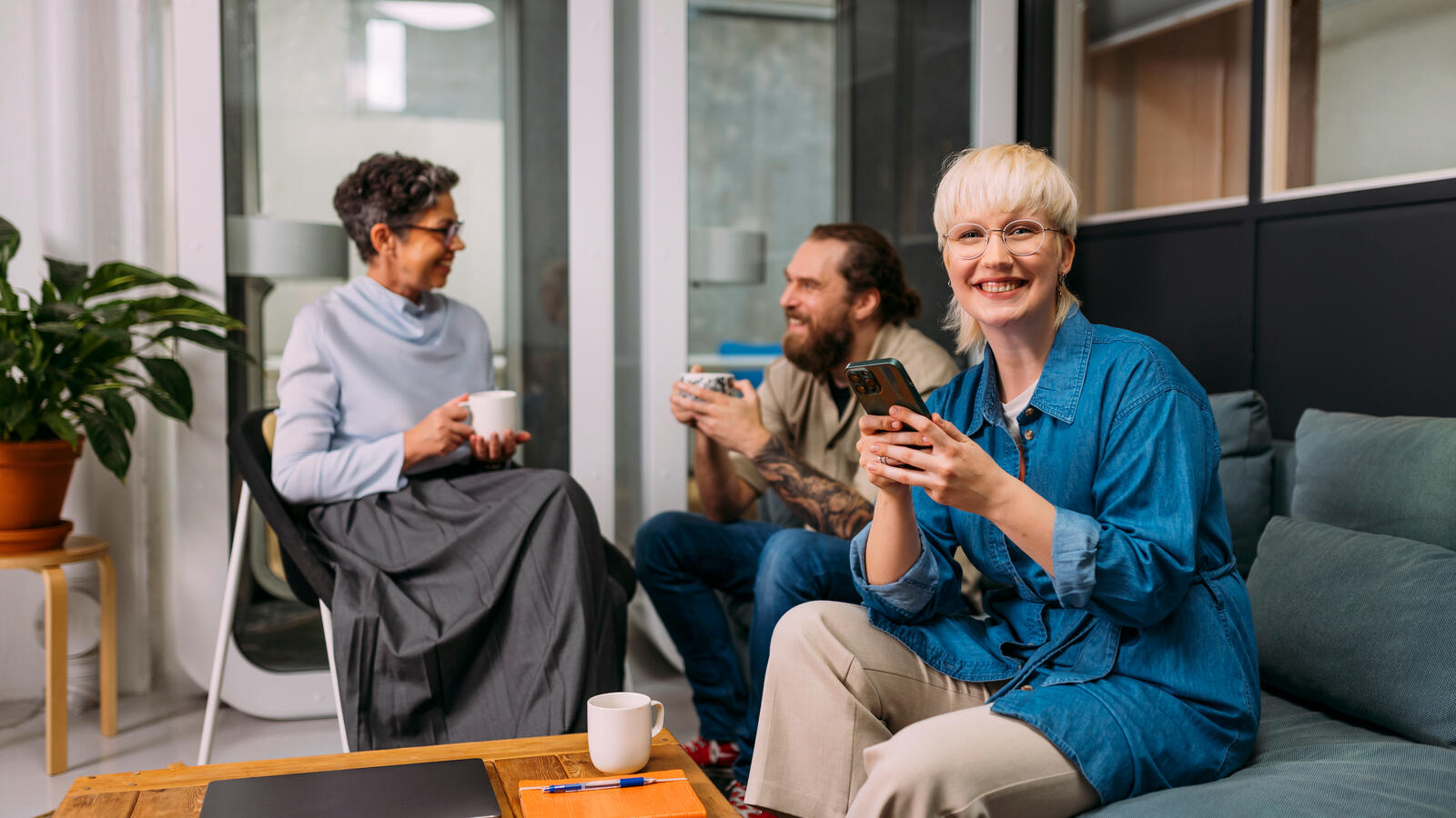 Kolme henkilöä istuu toimiston sohvaryhmässä, kahvikupit käsissä ja yksi katsoo puhelinta, pöydällä muistikirja ja kynä.