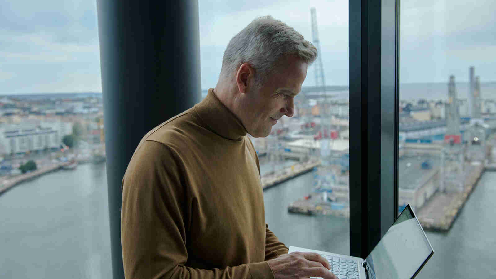 Mies seisoo ikkunan edessä kannettava tietokone käsissään, taustalla näkyy satama ja merimaisemaa.