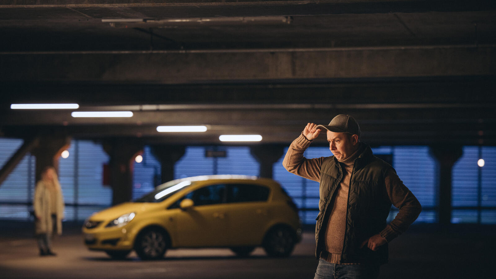 Mies seisoo mietteliään näköisenä ja pitää kiinni lippiksestään. Taustalla seisoo nainen auton vieressä.