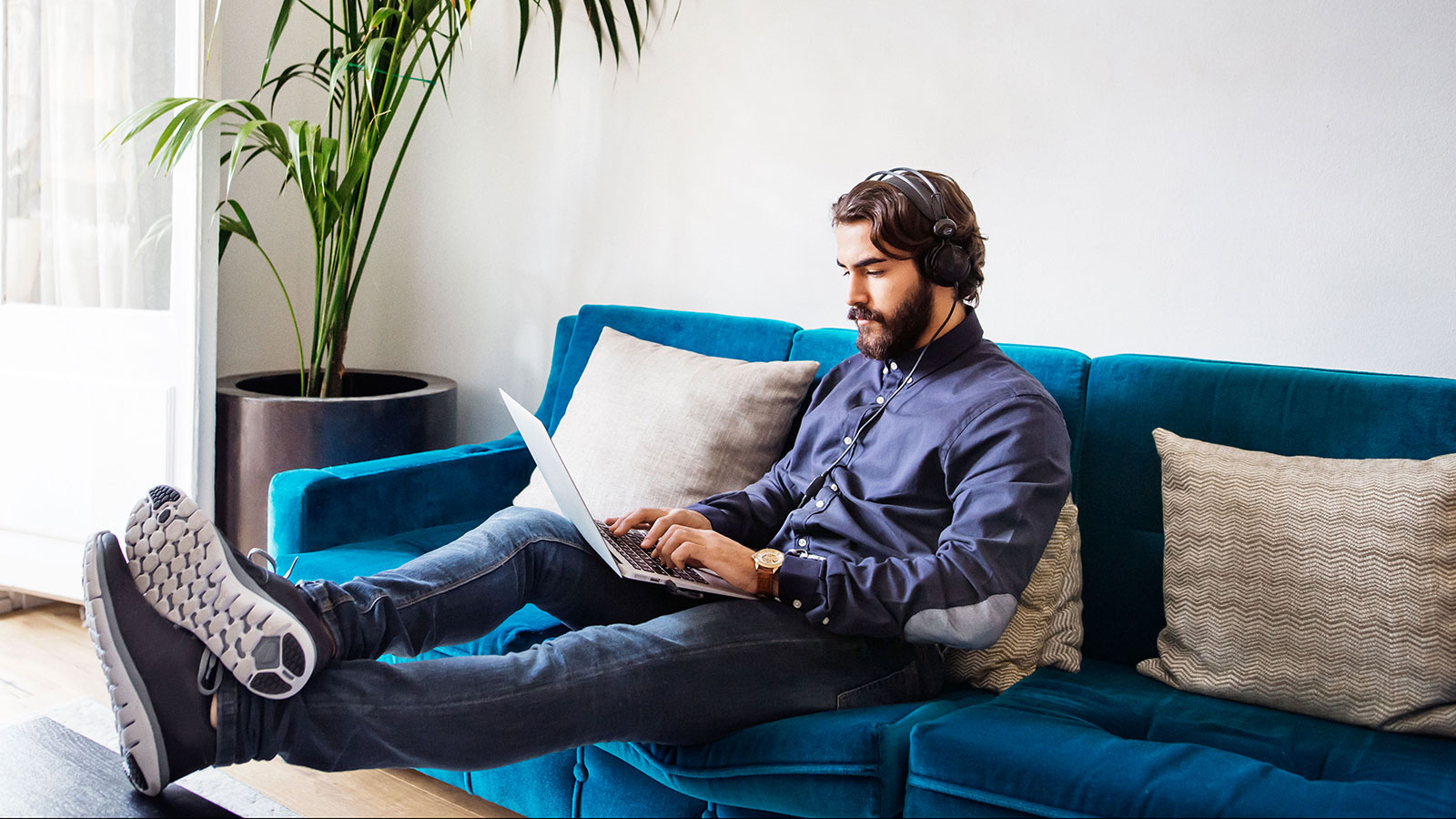 Tumma mies istuu sinisellä sohvalla läppäri sylissään.