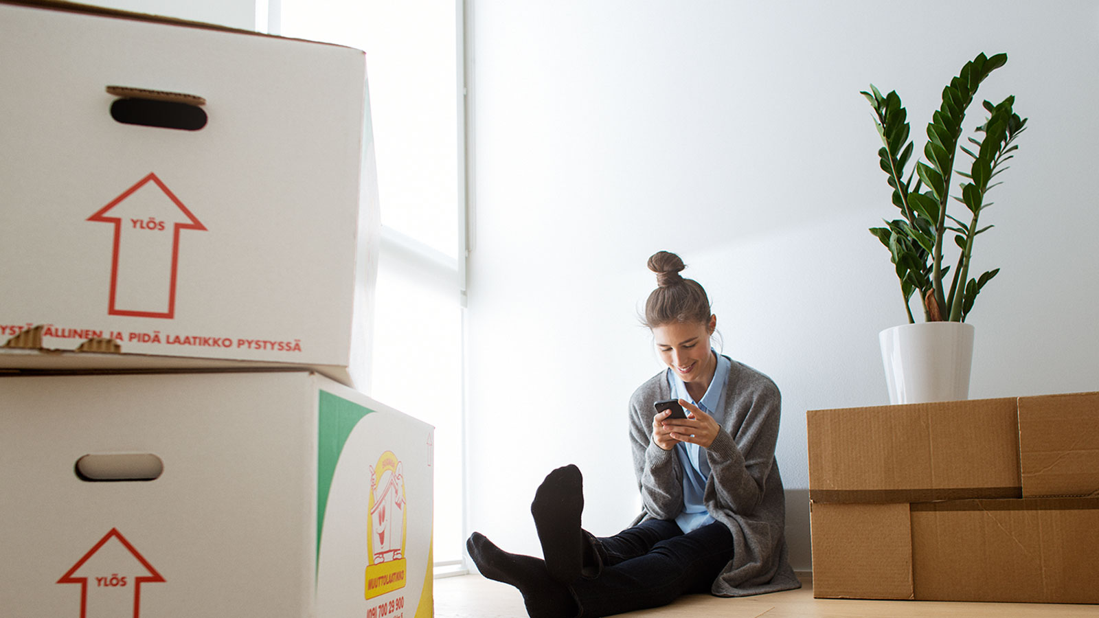 Nuori nainen istuu iloisena lattialla muuttolaatikoiden keskellä
