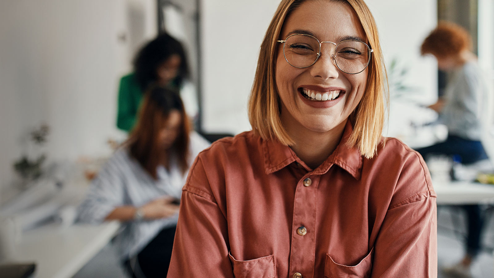 Silmälasipäinen nainen nauraa huolettomasti, tietäen että terveysturva ylläpitää työhyvinvointia.