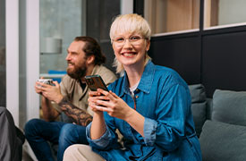 Kaksi henkilöä istuu modernissa toimistotilassa sohvalla ja käyttää älypuhelimia. Taustalla lasiseinä ja vaalea puupaneeli luovat nykyaikaisen työympäristön.