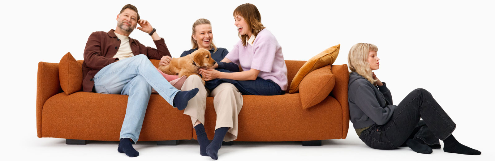 Valkoisessa ympäristössä oranssi sohva, jossa istuu mies, nainen ja tyttö. Sohvalla on myös koira, kultaisennoutajan pentu. Toinen tyttö istuu lattialla sohvan vieressä.