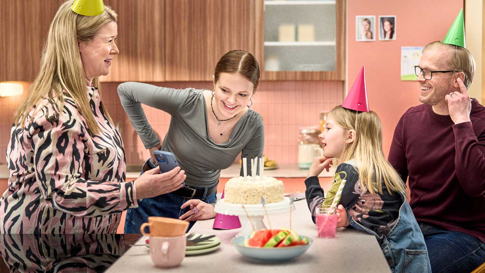 Ruokapöydän ääressä äiti, isä ja kaksi lasta. Pöydällä on täytekakku. Äiti näyttää kaikille jotain puhelimesta.