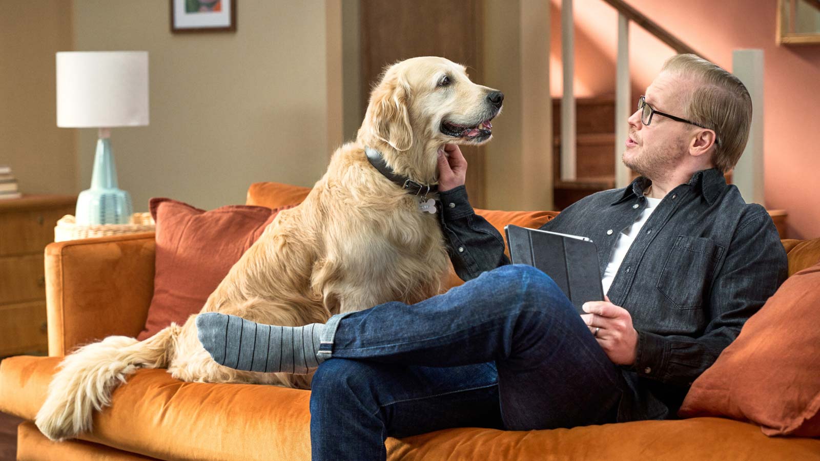 Mies ja koira istuvat oranssilla sohvalla.