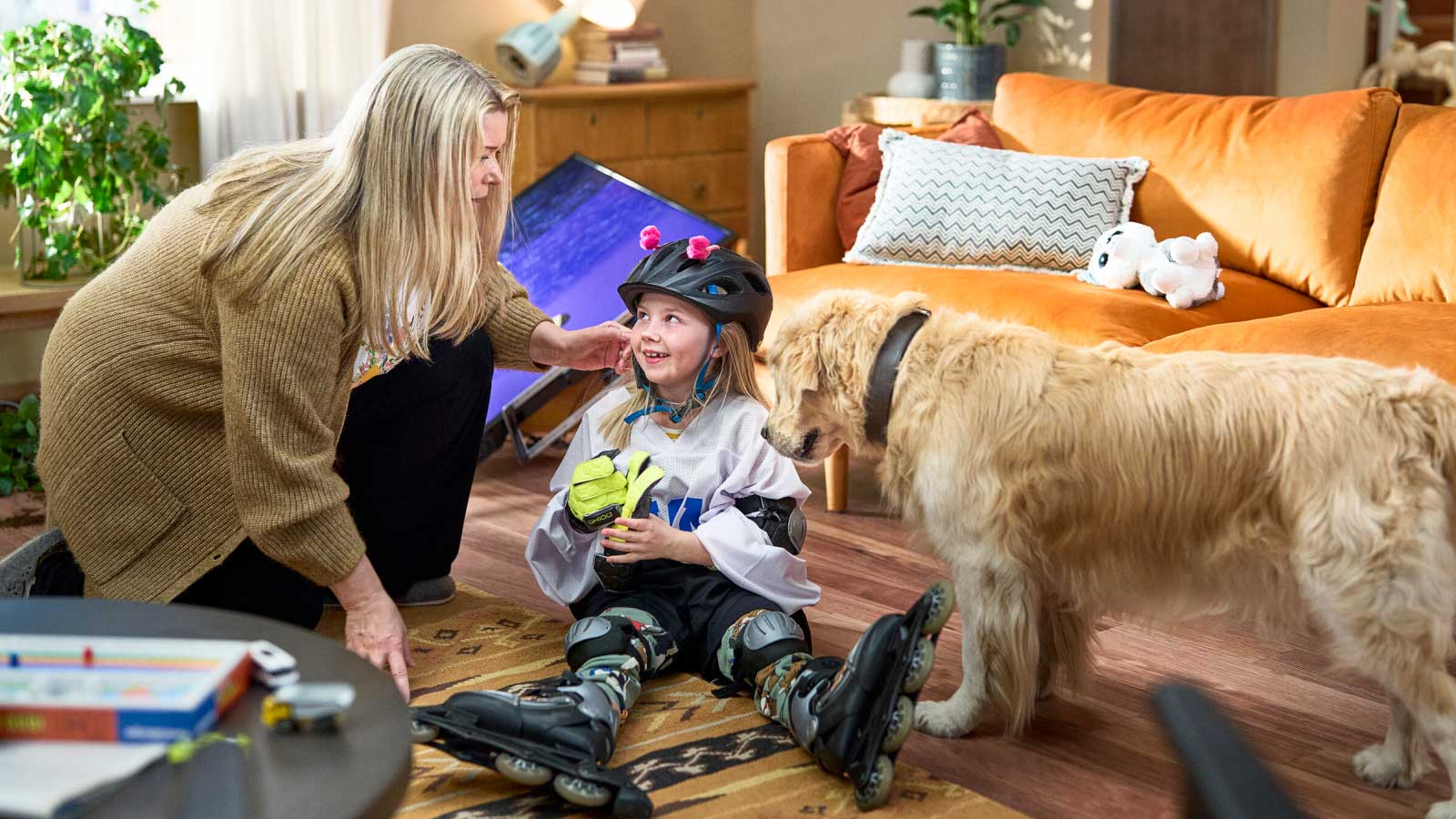 Lapsi istuu lattialla rullaluistimet jalassa. Taustalla on kaatunut taulutelevisio. Äiti on kyykistynyt lapsen viereen, lapsi hymyilee ja perheen koira, kultainennoutaja katsoo myös lasta. 