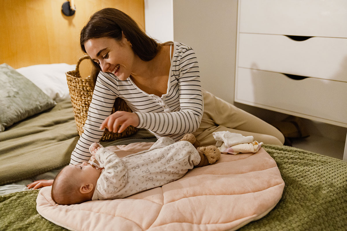 En leende mamma småpratar med en bebis som ligger på sängen.