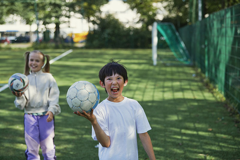 Två barn ler skälmskt med fotbollar i händerna på en spelplan.